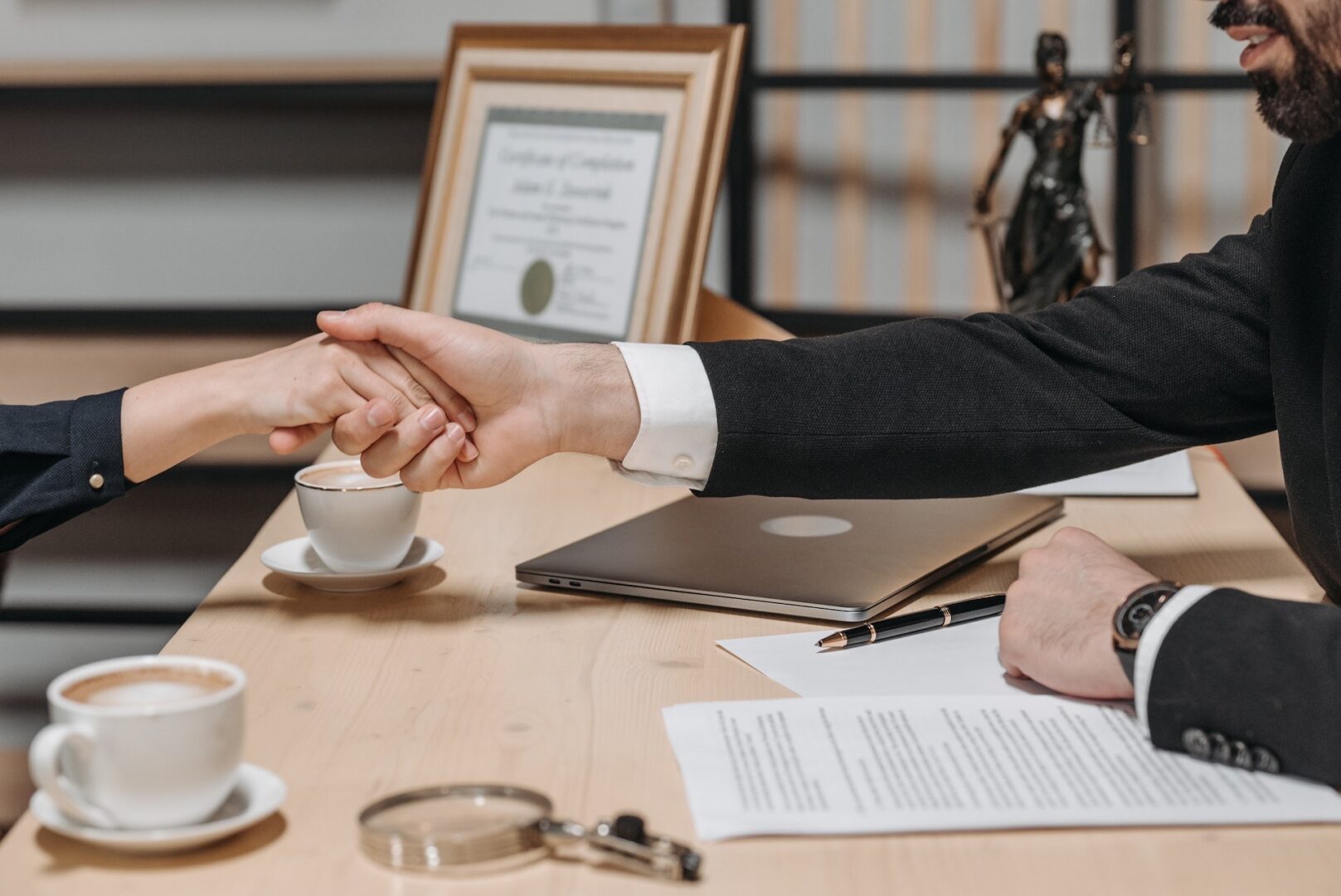 Two people shaking hands over a table with papers and coffee.